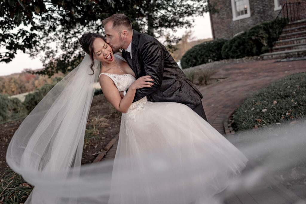 bride and groom doing a dip with veil flying towards camera. Bride laughing while groom kissing bride on cheek