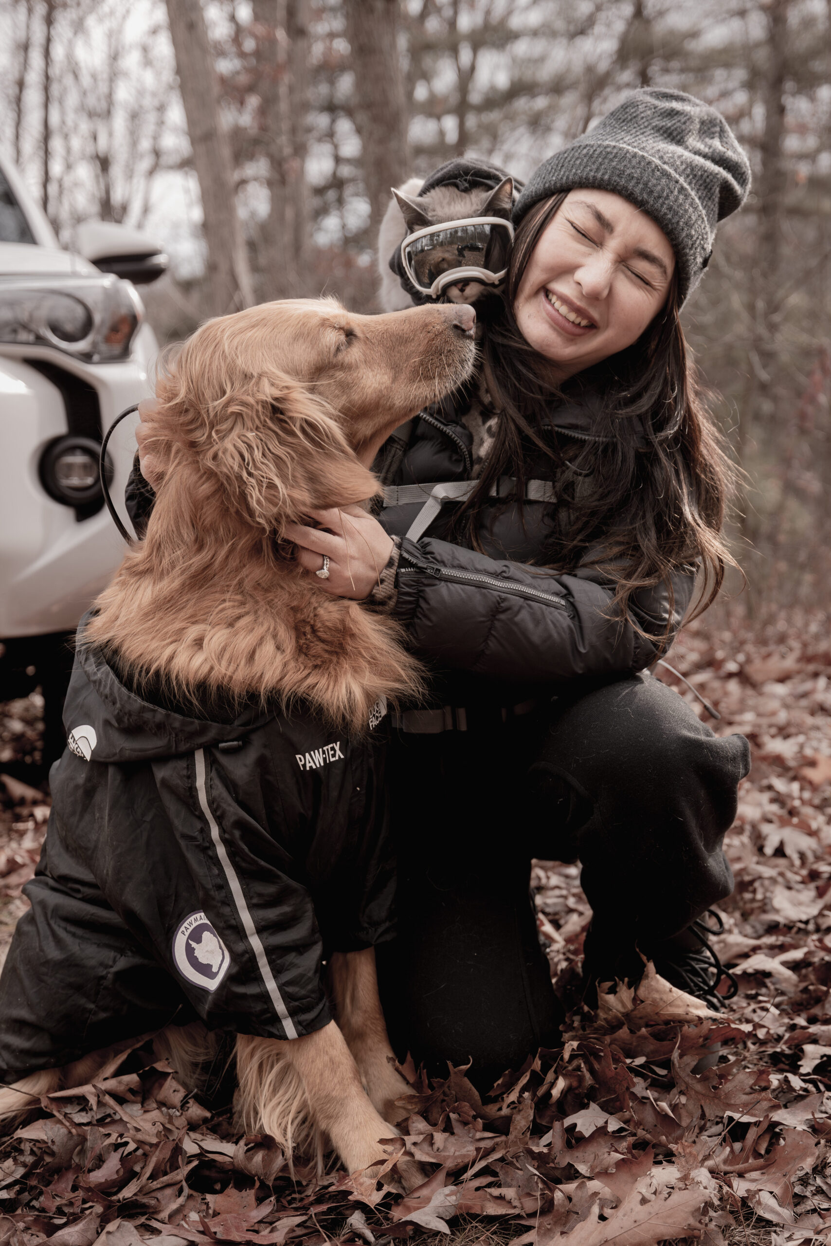 Girl dressed in winter camping attire, camping with her pet cat in tiny cat goggles and her red golden retriever kissing her on the cheek.