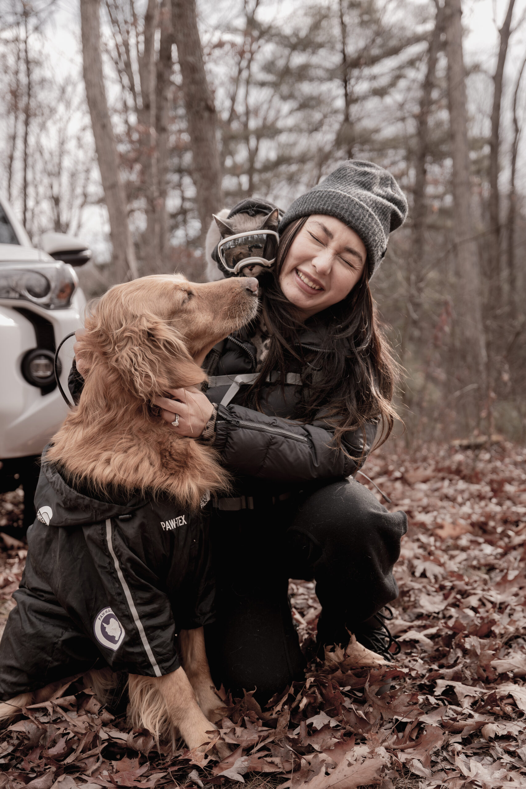 Girl dressed in winter camping attire, camping with her pet cat in tiny cat goggles and her red golden retriever kissing her on the cheek.