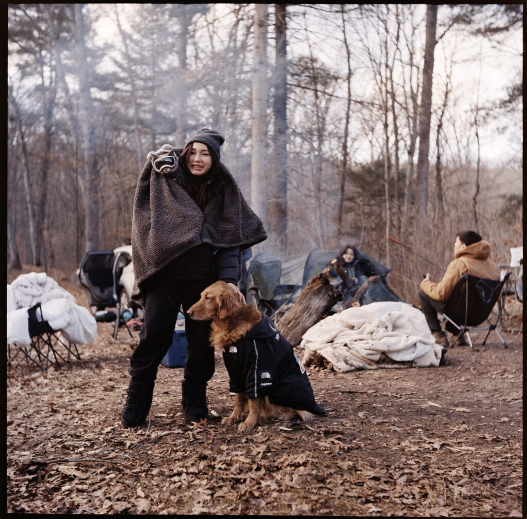 girl in blanket on campsite with her cat on her shoulder and red golden retriever next to her smiling.