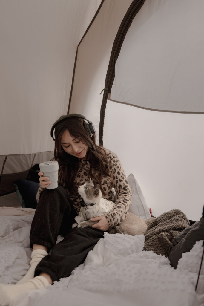 Woman sitting in tent with her cat with Bang and Olufsen over ear headphones on drinking coffee on blankets.